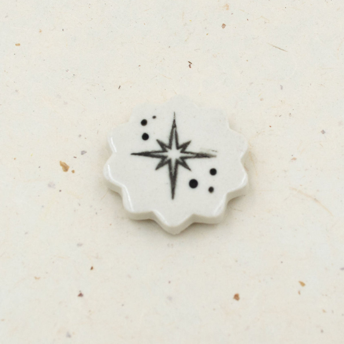 A small white ceramic or porcelain star-shaped tile with a black starburst and dot pattern, placed on a light beige textured surface.