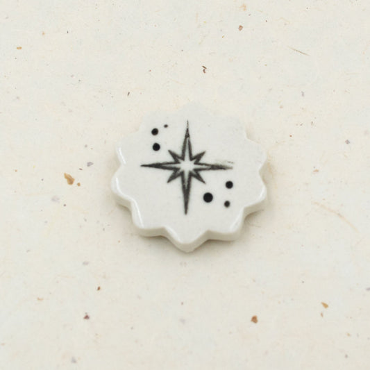 A small white ceramic or porcelain star-shaped tile with a black starburst and dot pattern, placed on a light beige textured surface.